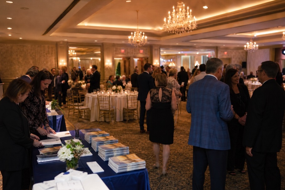 A wide, warmly lit ballroom set up for a nonprofit fundraising gala. Round tables with white tablecloths and simple floral centerpieces fill the space while guests in business casual and formal attire stand and mingle between them. In the foreground, a registration table displays neatly stacked printed programs and materials. Chandeliers and soft overhead lighting create a polished, welcoming atmosphere with a shallow depth of field that keeps the background softly blurred.